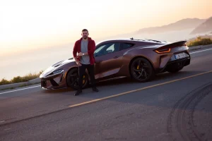 A modern car enthusiast standing confidently beside a sleek, high-performance sports car on a scenic coastal highway at golden hour, with dynamic lighting reflecting off the polished paint, distant mountains and ocean in the background, subtle tire marks on the asphalt suggesting motion, and a sense of freedom and passion for driving
