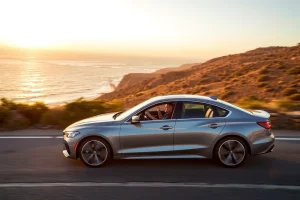 A sleek modern car on a winding coastal road at sunset, its new alloy wheels gleaming, with a relaxed driver seated in a luxurious interior, the cabin visible through a panoramic glass roof, showcasing comfort and pleasure of driving