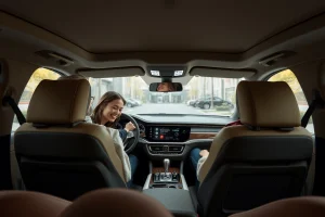 A modern, comfortable car interior with a focus on ergonomic seating, soft ambient lighting, and a sleek dashboard featuring the koleso-bestune logo subtly integrated into the steering wheel or center console. The scene conveys relaxation and premium comfort, with a driver and passenger smiling gently, wearing casual clothing, in a quiet urban or suburban setting visible through the windows. Warm, natural tones dominate — beige, soft gray, and warm wood accents — enhancing the feeling of luxury