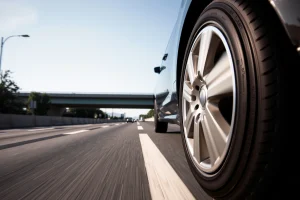 A modern city street with a car driving smoothly, showing close-up of alloy wheels on the vehicle, with a highway in the background under clear daylight, emphasizing wheel design, tire tread, and urban-road transition, realistic lighting, no text or logos