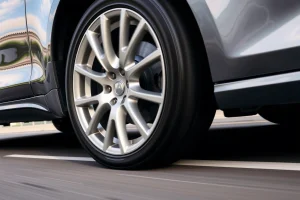 A close-up of lightweight yet durable alloy wheels on a modern car, showing their sleek design, strong spokes, and reflective metallic surface, with a blurred urban highway background suggesting motion and performance
